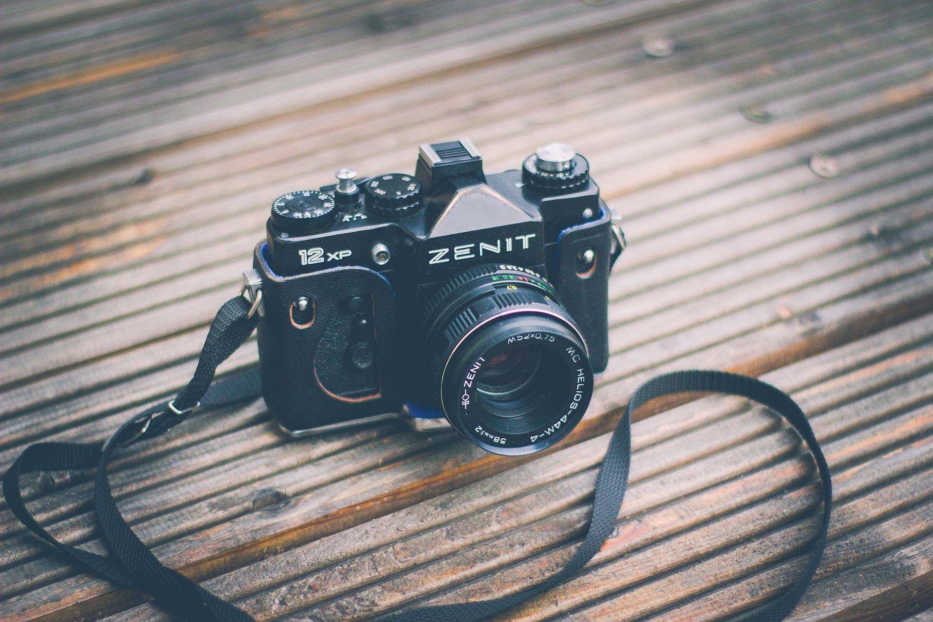 A black Zenit-12XP film camera fitted with a lens, resting on a wooden surface